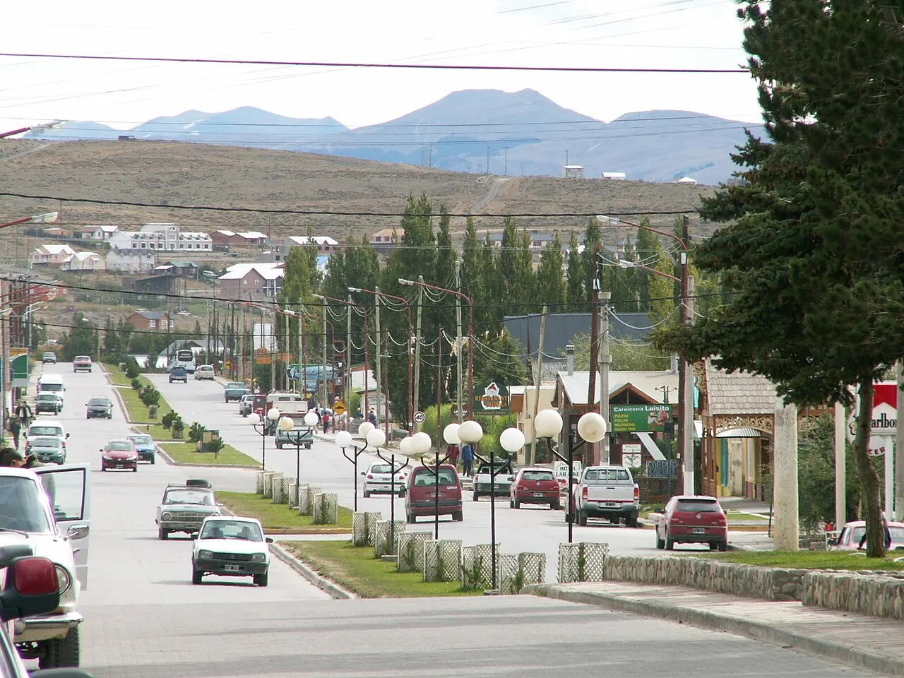 A Avenida del Libertador, em El Calafate, com carros, postes, árvores no canteiro central e colinas secas ao fundo.