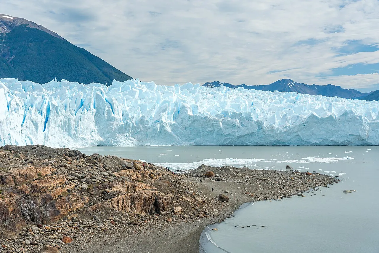 A frente do Glaciar Perito Moreno, uma parede de gelo azulado e rachado sobre a água do lago, vista de uma margem de pedras.