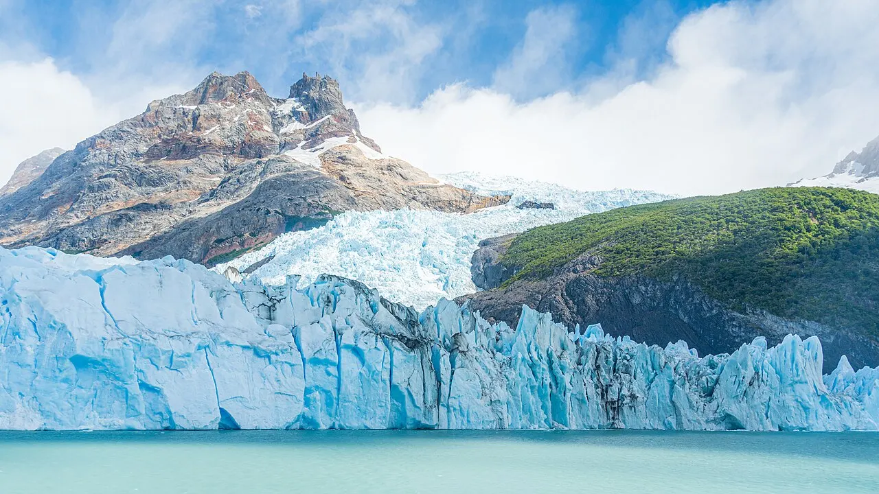 A frente do Glaciar Spegazzini, uma parede de gelo branco e azul sobre o lago, com uma encosta verde e picos rochosos atrás.