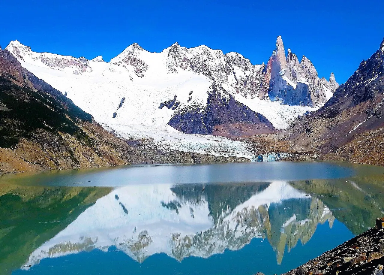 A Laguna Torre com a água parada refletindo o Cerro Torre e as montanhas nevadas, com uma geleira descendo até o lago.