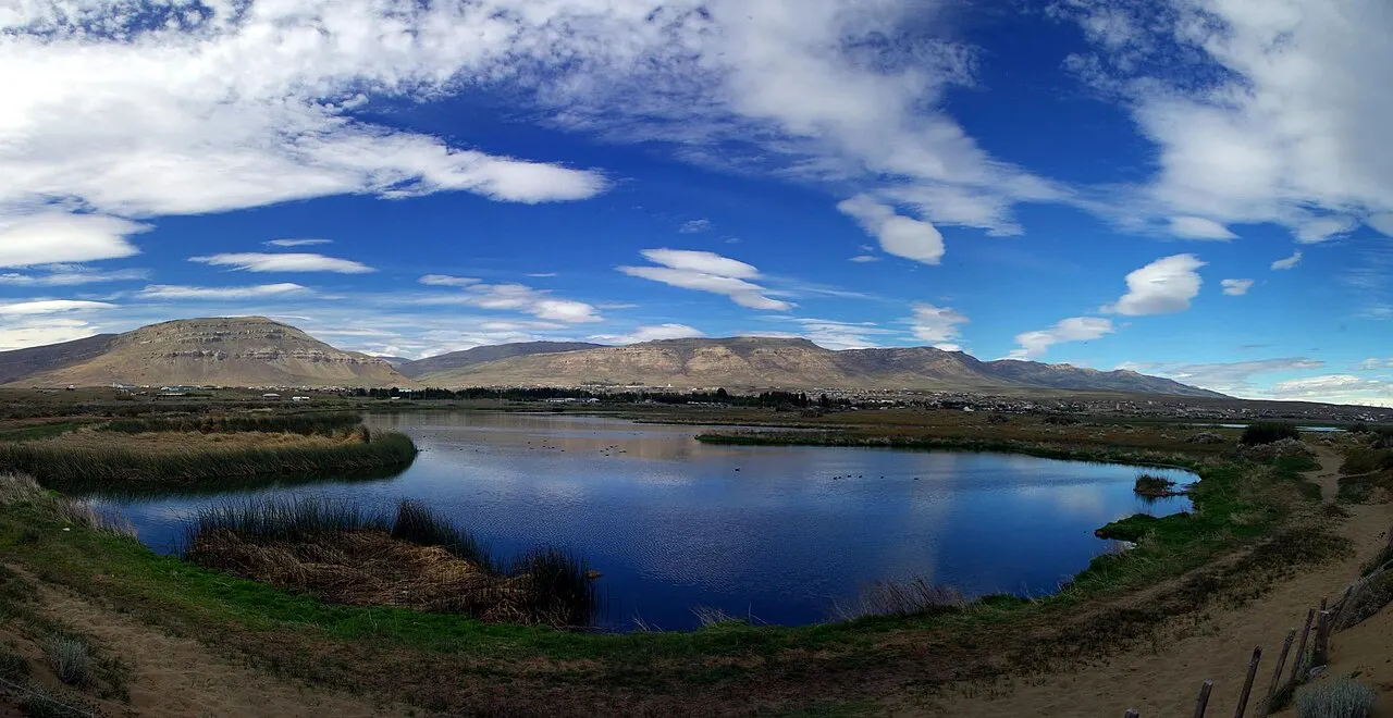 A Laguna Nimez, uma lagoa azul cercada de capim, com colinas da estepe ao fundo e céu com nuvens.