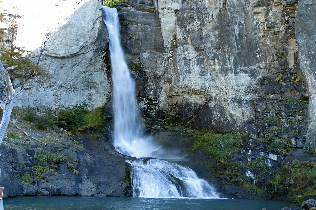 Cascata branca que cai de uma parede alta de rocha cinza em dois saltos até um poço de água, com árvores e musgo nas laterais.