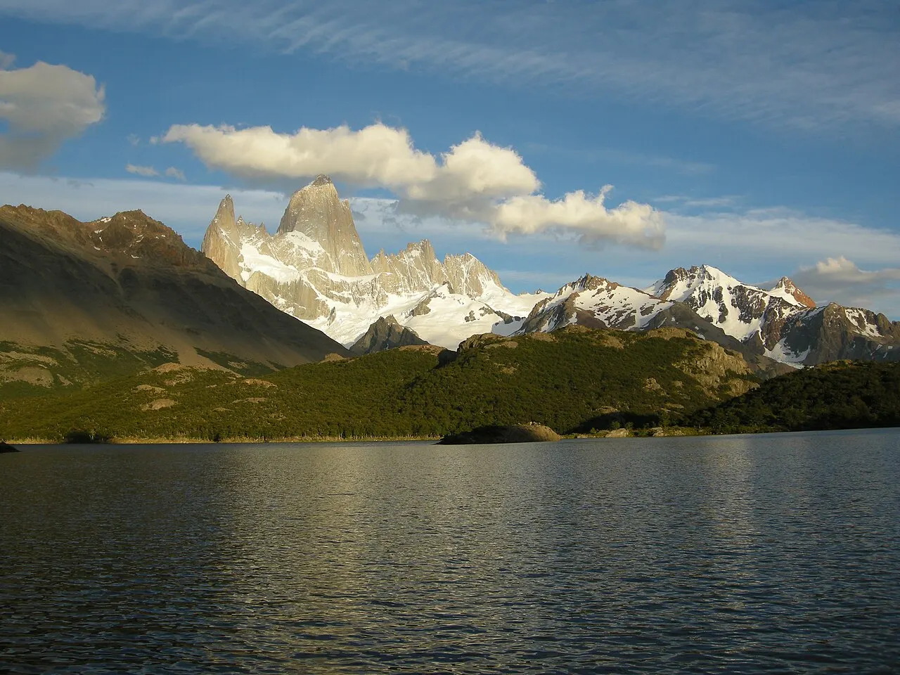 Lagoa escura e calma em primeiro plano, uma faixa de floresta verde na outra margem e, acima dela, o maciço do Fitz Roy com neve, sob nuvens brancas.