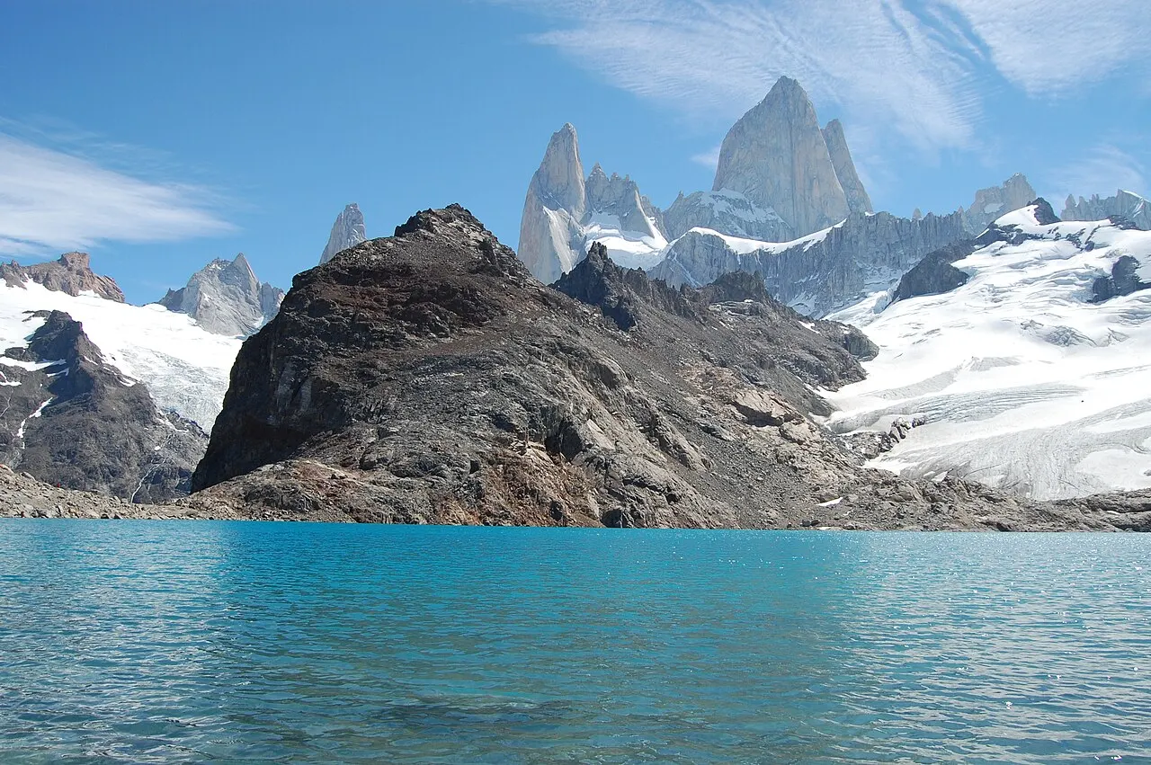 Lagoa de água azul-turquesa em primeiro plano, um morro escuro de rocha atrás e, ao fundo, as agulhas de granito do Fitz Roy com geleiras e neve sob céu azul.