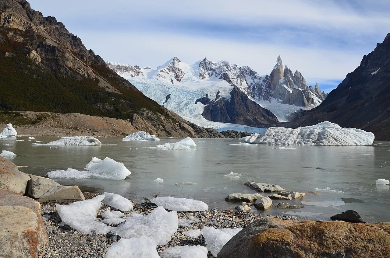 Lagoa cinzenta com blocos de gelo flutuando e pedaços de gelo na margem de pedras; ao fundo, a geleira desce entre encostas escuras até as agulhas nevadas do Cerro Torre.