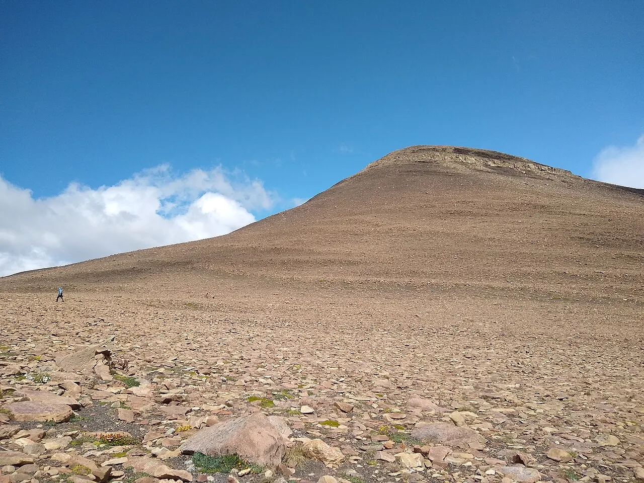 Encosta larga e arredondada de cascalho e pedras soltas subindo até o cume, sob céu azul com nuvens brancas; uma pessoa minúscula caminha ao longe à esquerda.