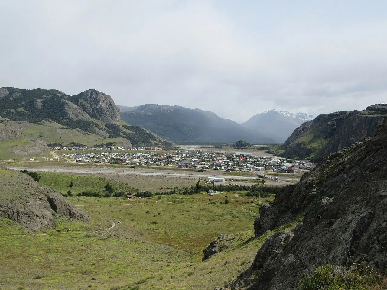 Vista do alto de uma encosta rochosa com capim verde: a vila de El Chaltén espalhada no fundo do vale, entre montanhas, sob céu nublado.