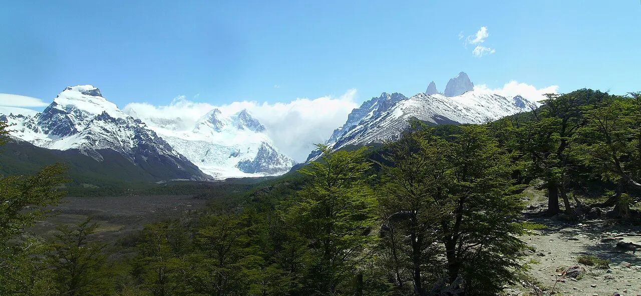 Panorama com árvores verdes em primeiro plano, o vale amplo abaixo e uma fileira de montanhas nevadas ao fundo, com nuvens presas aos cumes.