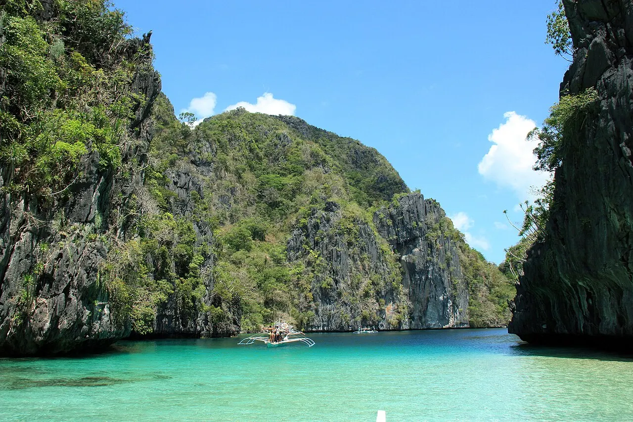 Shallow pale-green water between two forested limestone walls, with an outrigger boat in the distance under a blue sky.