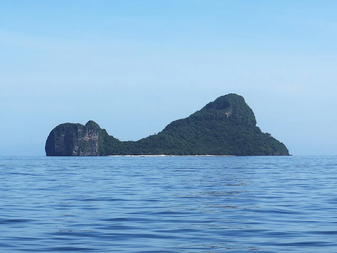 Forested limestone island seen from afar, with a low rock at one end, a strip of pale sand at its base and calm sea in the foreground.