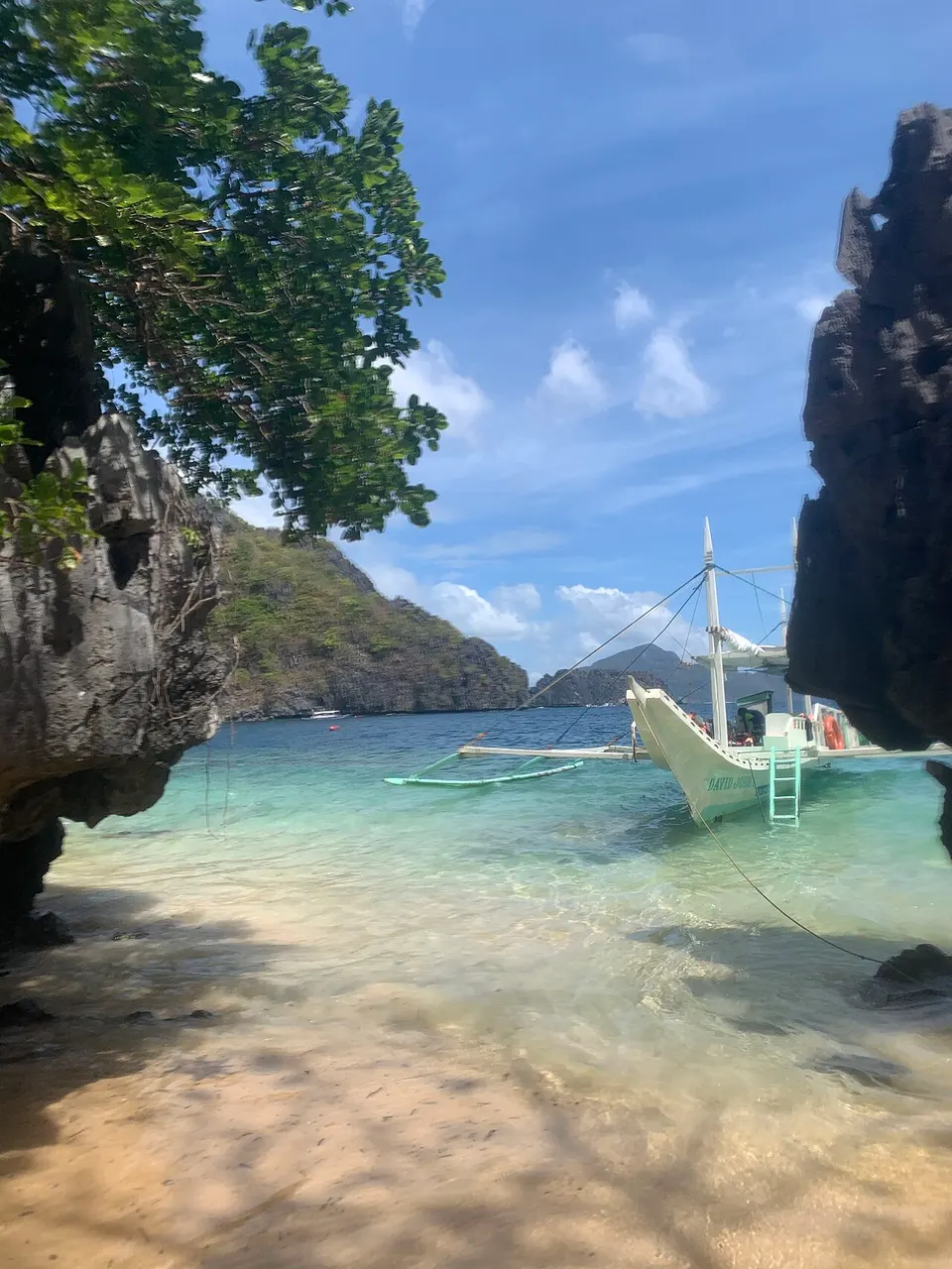 Shaded sand and clear shallow water between two dark limestone rocks, with an anchored outrigger boat and a hill in the background.
