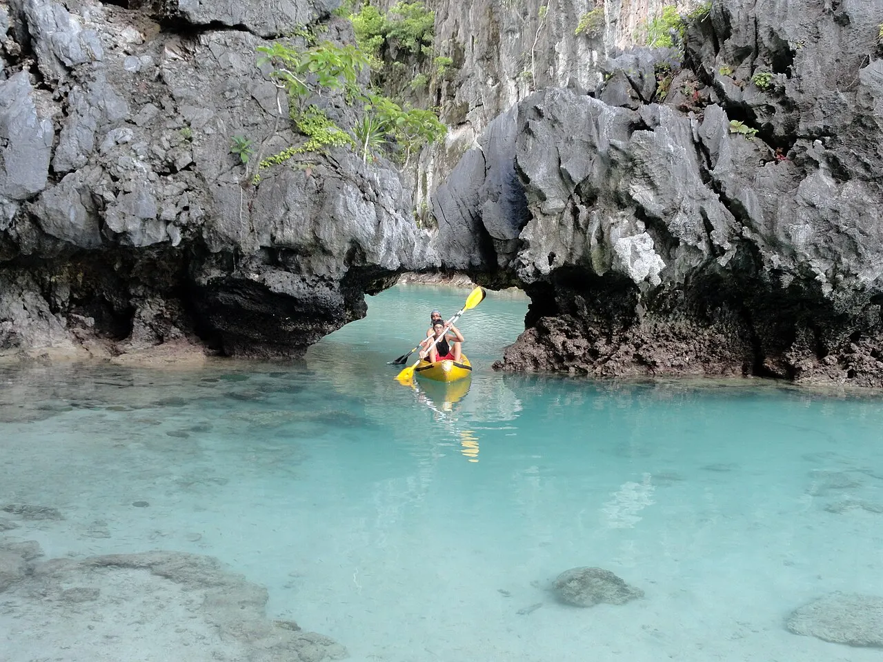 Yellow kayak with two people passing through an opening between limestone rocks, in a lagoon of clear turquoise water with stones on the bottom.