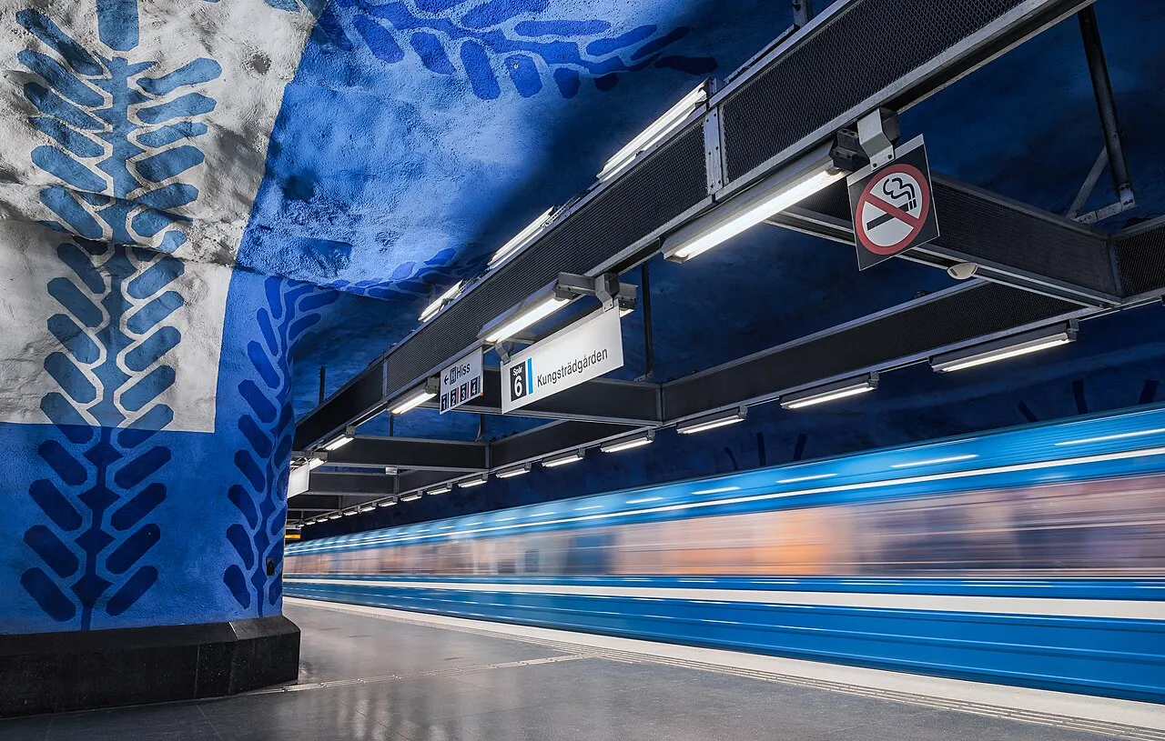 A parede de rocha da estação está pintada de azul com formas brancas, e um trem azul passa embaçado junto à plataforma.