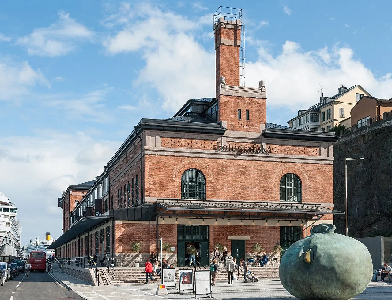 Edifício de tijolo do Fotografiska em Estocolmo, com chaminé alta, o nome na parede e pessoas pequenas na calçada.