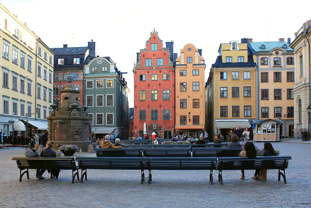 Place historique entourée de façades colorées à pignons à degrés, avec une fontaine en pierre au centre et des gens assis sur des bancs sur les pavés.