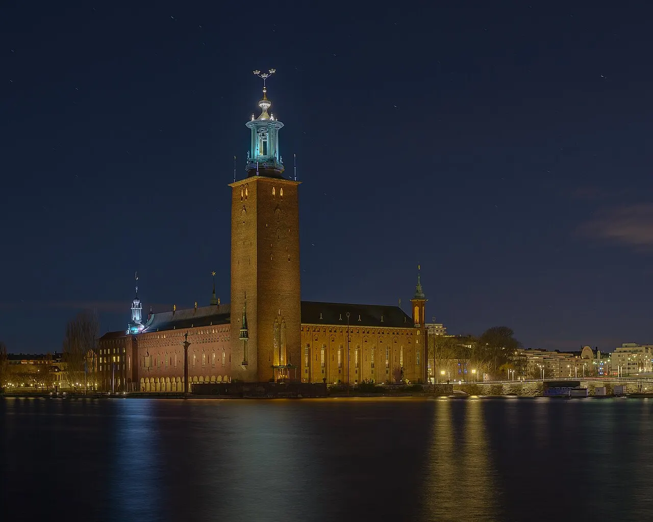 Prefeitura de Estocolmo iluminada à noite, com a torre alta de tijolo e a coroa dourada no topo, vista da água.
