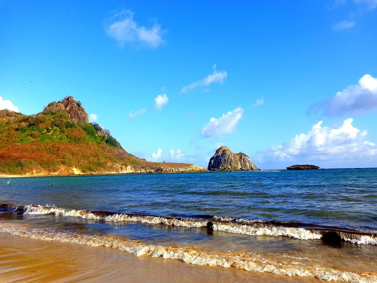 A Baía do Sueste, com ondas pequenas na areia, mar azul e, ao fundo, um morro de encosta avermelhada e ilhotas de pedra.