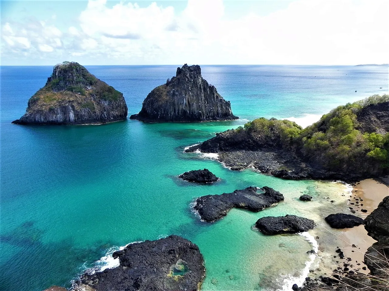A Baía dos Porcos vista de cima, com piscinas de água turquesa entre pedras escuras e os dois rochedos do Morro Dois Irmãos no mar.