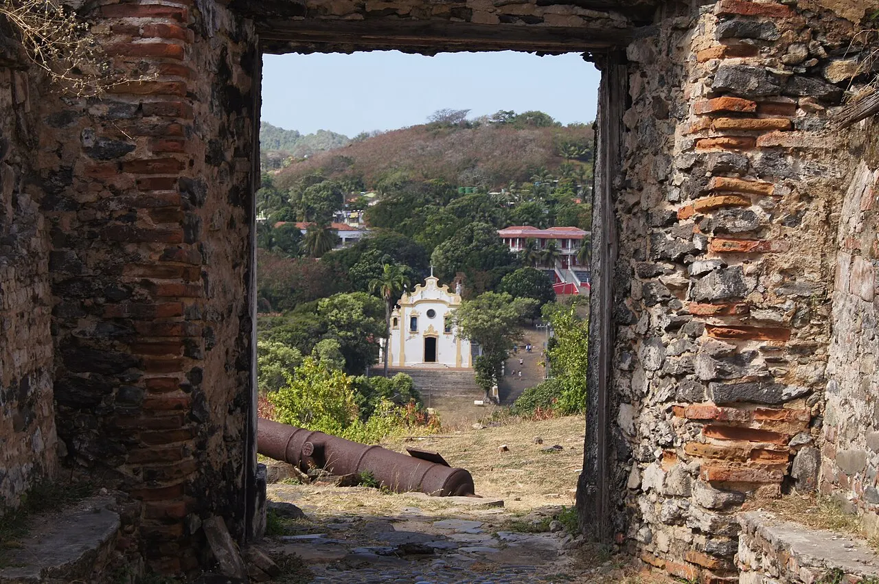 Um portal de pedra e tijolo do Forte de Nossa Senhora dos Remédios, com um canhão antigo no chão e, ao fundo, a igreja branca da vila entre árvores.