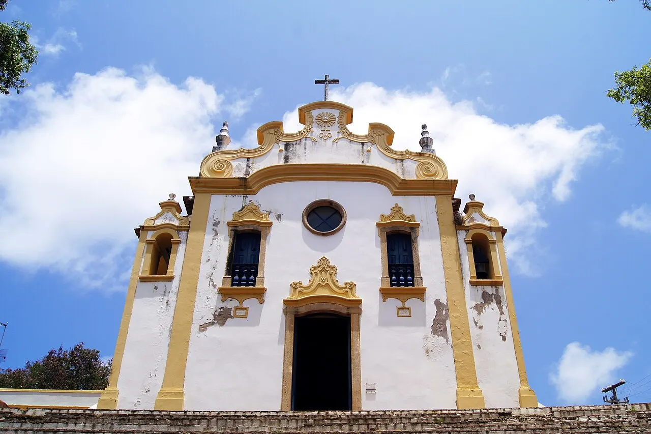 A fachada da Igreja de Nossa Senhora dos Remédios, branca com molduras amarelas, uma cruz no topo e janelas azuis, vista de baixo contra o céu azul.