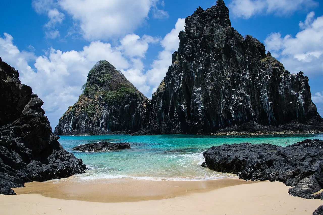 Um trecho de areia clara da Praia da Cacimba do Padre entre pedras escuras, com água turquesa e o paredão do Morro Dois Irmãos sob céu azul com nuvens.