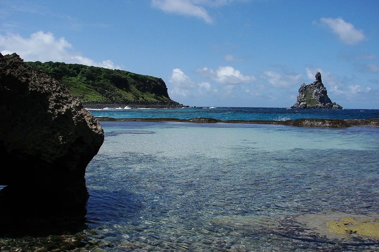 A piscina natural da Praia do Atalaia, com água rasa e transparente sobre o fundo de pedra, uma rocha escura à esquerda e a Ilha do Frade no horizonte.