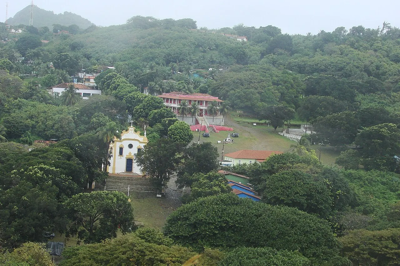 A Vila dos Remédios vista de cima, com a igreja branca e amarela e casas de telhado vermelho espalhadas entre árvores numa encosta verde, sob céu nublado.