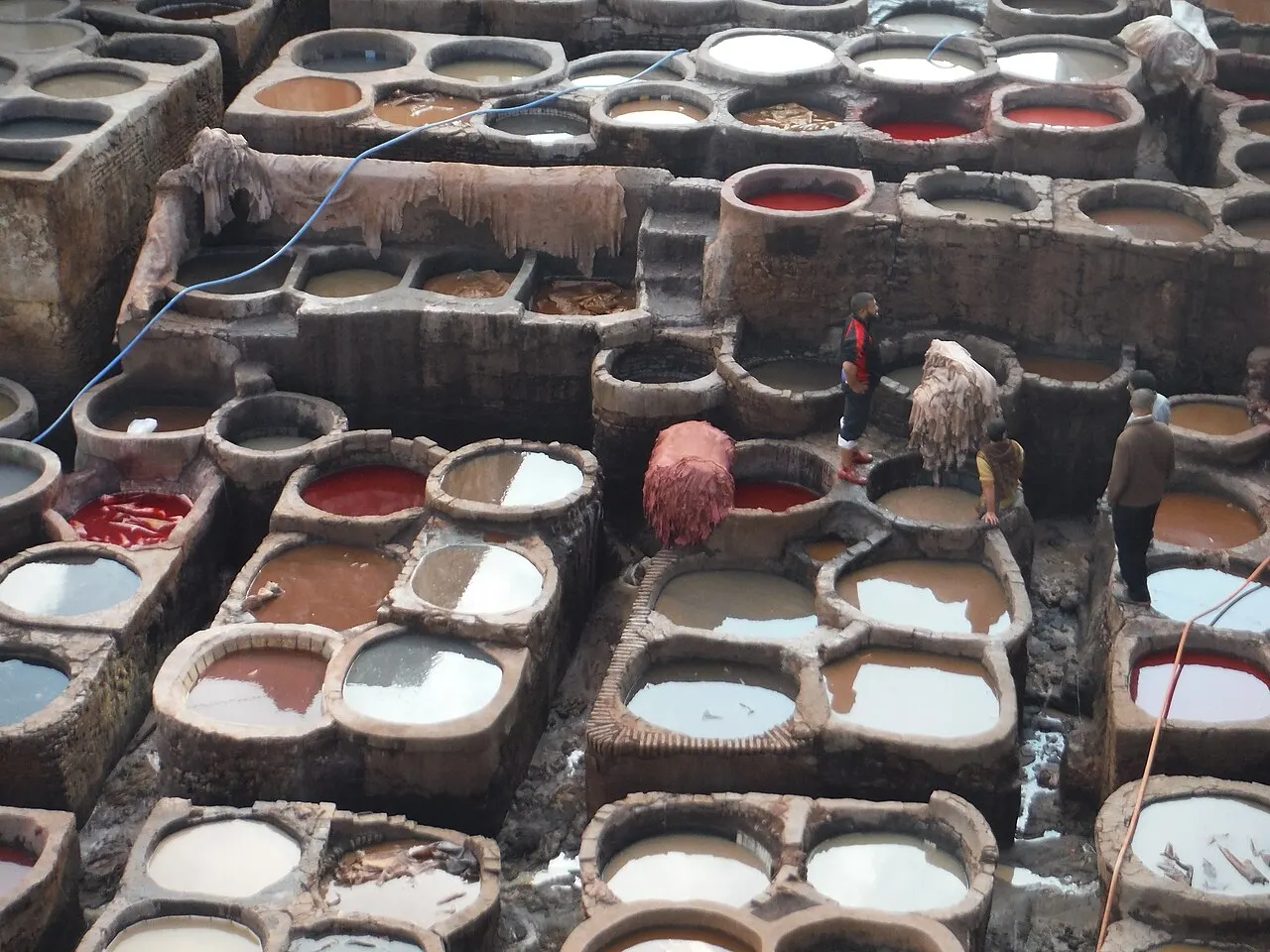 Vista de cima de dezenas de tanques redondos e retangulares de pedra, cheios de líquidos de cores diferentes como branco, vermelho e marrom, com peles penduradas e alguns trabalhadores caminhando entre os tanques.