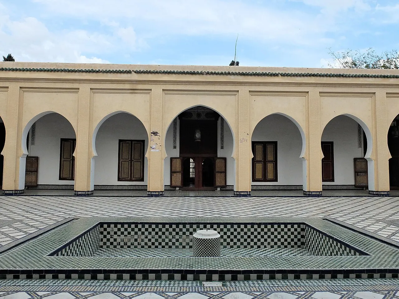 Uma arcada creme de arcos e portas de madeira fecha o pátio, com um tanque de azulejos em primeiro plano.