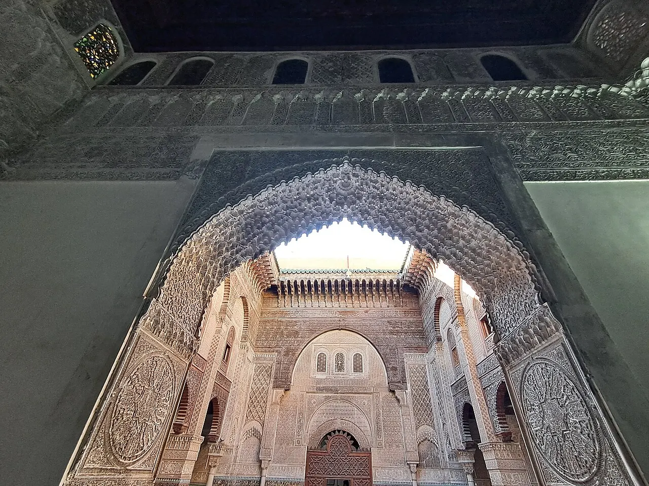 Um arco de entrada ricamente esculpido em gesso, com padrões geométricos entrelaçados, emoldurando a vista de um pátio interno decorado com mosaicos e talha de madeira, luz entrando por uma claraboia.