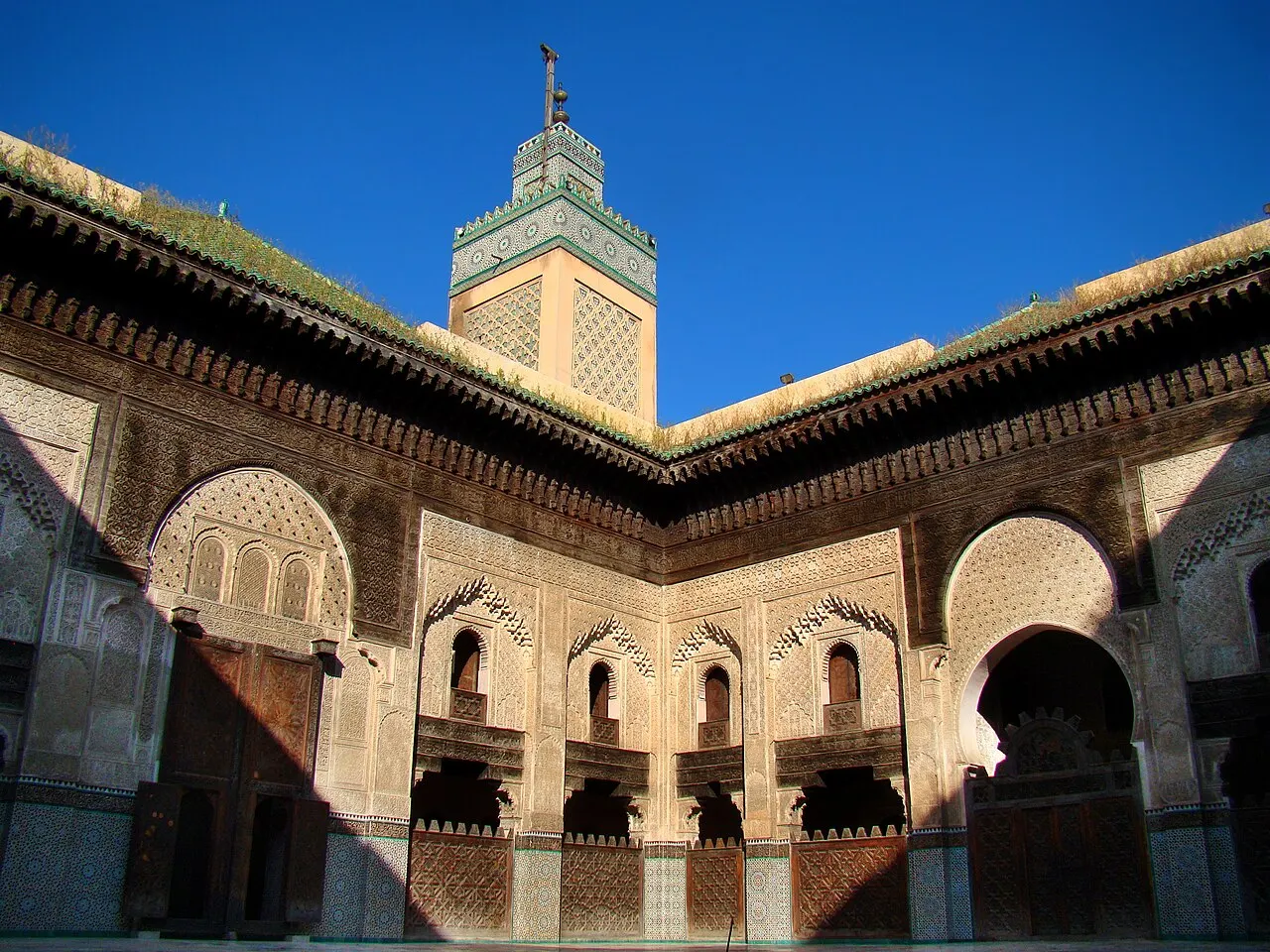 Um pátio interno cercado por paredes cobertas de talha geométrica detalhada em gesso e madeira, arcos ornamentados e um minarete quadrado ao fundo, sob céu azul intenso.