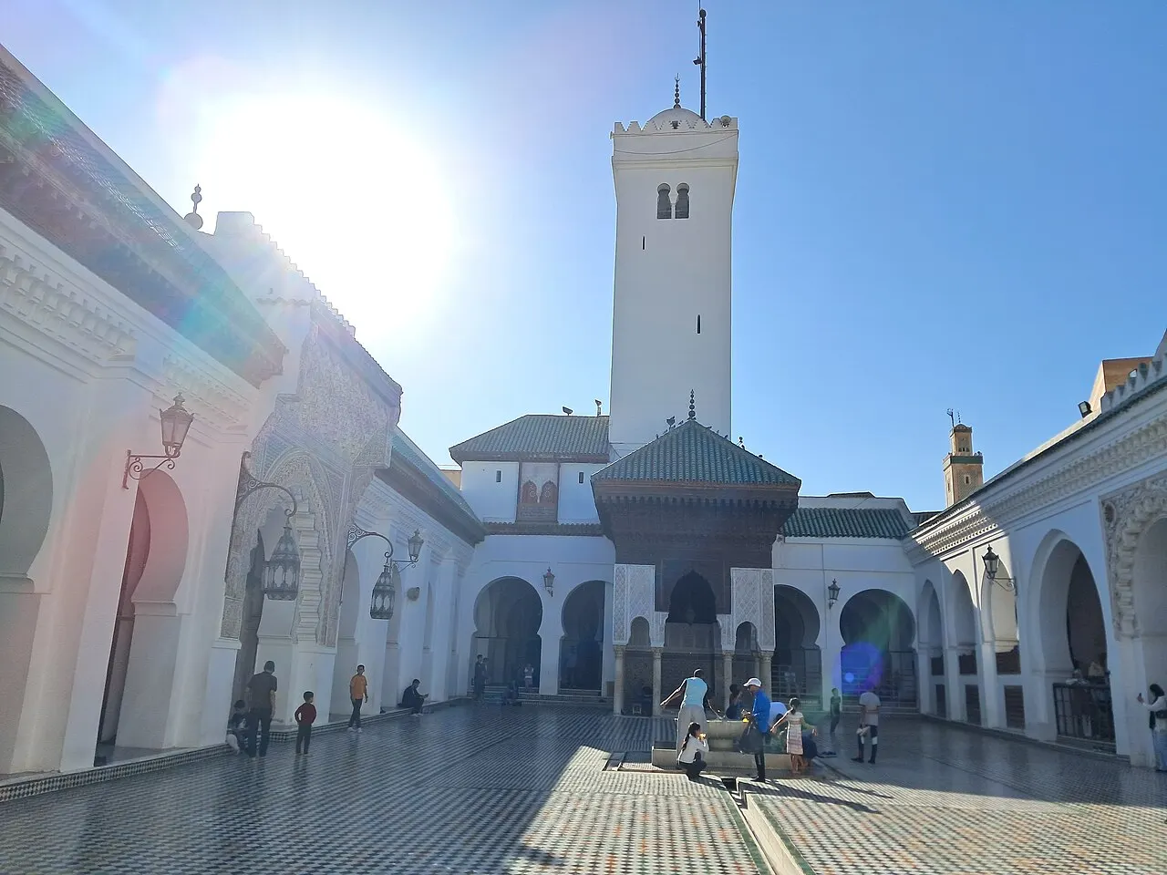 O pátio branco tem arcos, um minarete quadrado e um telhado verde no centro, com algumas pessoas e sol forte.
