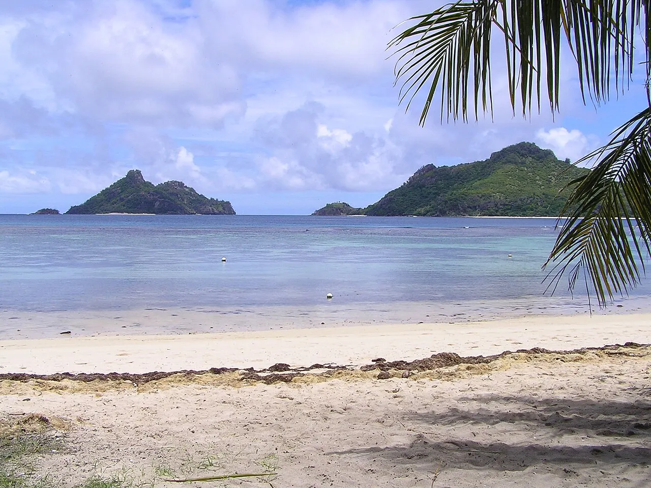 Une plage de sable clair avec des palmes de cocotier au premier plan, un lagon peu profond bleu clair et deux îles hautes et vertes à l'horizon.