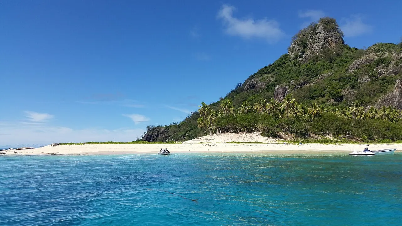 Une île avec une plage de sable blanc, des cocotiers et une colline boisée qui finit en pic rocheux, vue depuis la mer turquoise, avec deux scooters des mers et un bateau près du rivage.