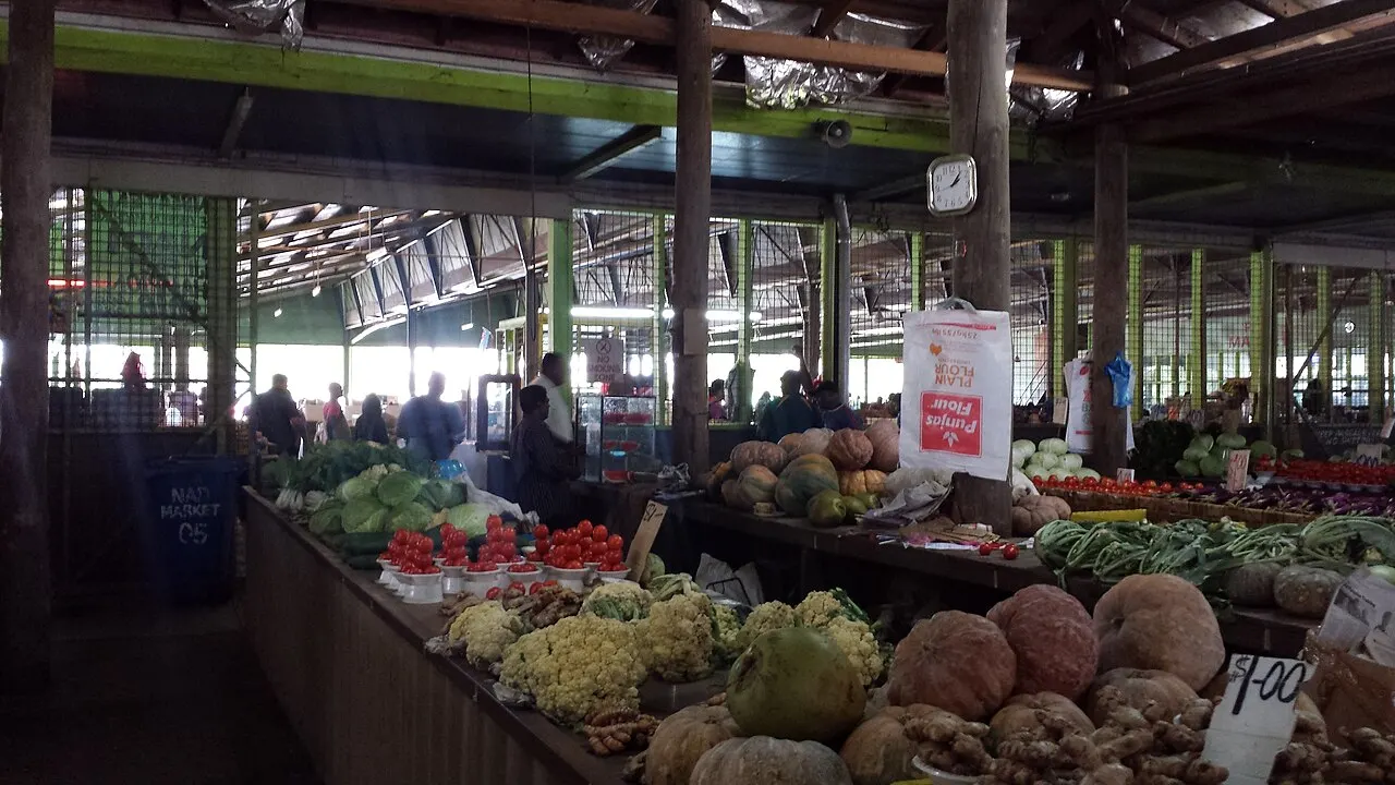 L'intérieur couvert d'un marché, avec des étals de courges, de choux-fleurs, de gingembre, de tomates et de haricots rangés en tas, et des gens au fond entre les piliers.