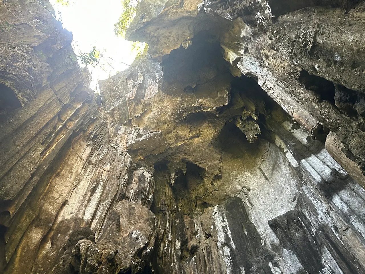 Les parois et le plafond calcaires d'une grotte vus d'en bas, avec la roche en couches et la lumière du jour qui entre par une ouverture en hauteur.