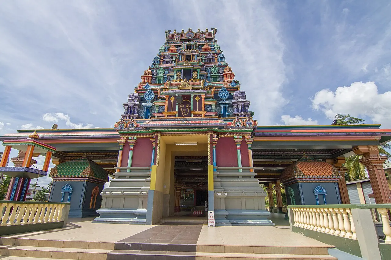 Temple hindou à la tour en pyramide couverte de sculptures peintes de nombreuses couleurs, au-dessus d'une entrée à colonnes, avec un escalier et des balustrades claires devant, sous un ciel bleu nuageux.