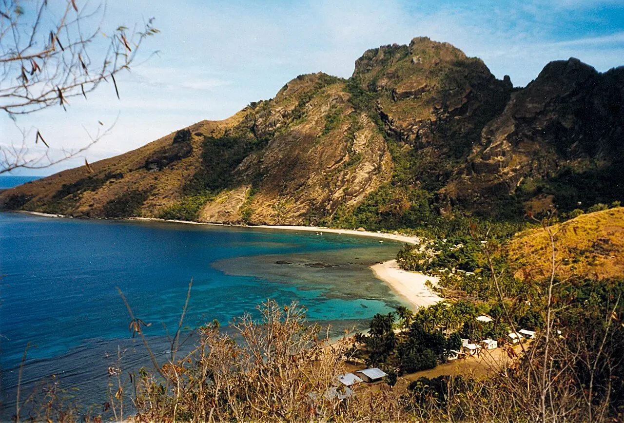 Une baie vue d'en haut, avec une eau bleue, un récif peu profond près du rivage, une bande de sable blanc et des maisons basses entre les arbres, au pied de montagnes de roche sombre.