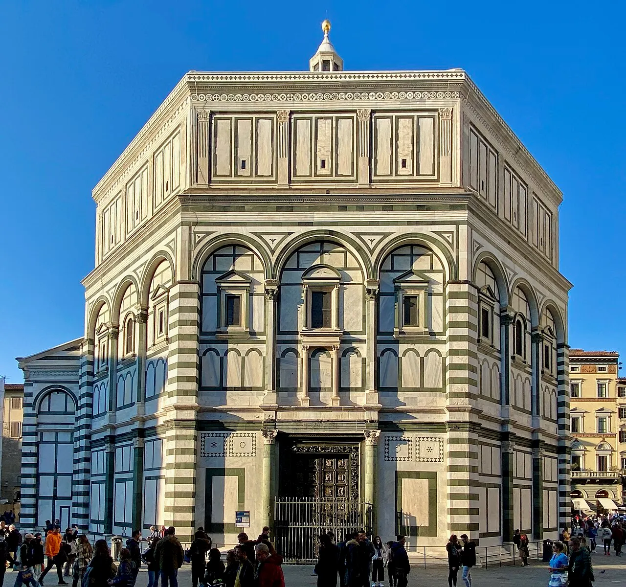 Baptistère octogonal de marbre blanc et vert à arcades, sous un ciel bleu.