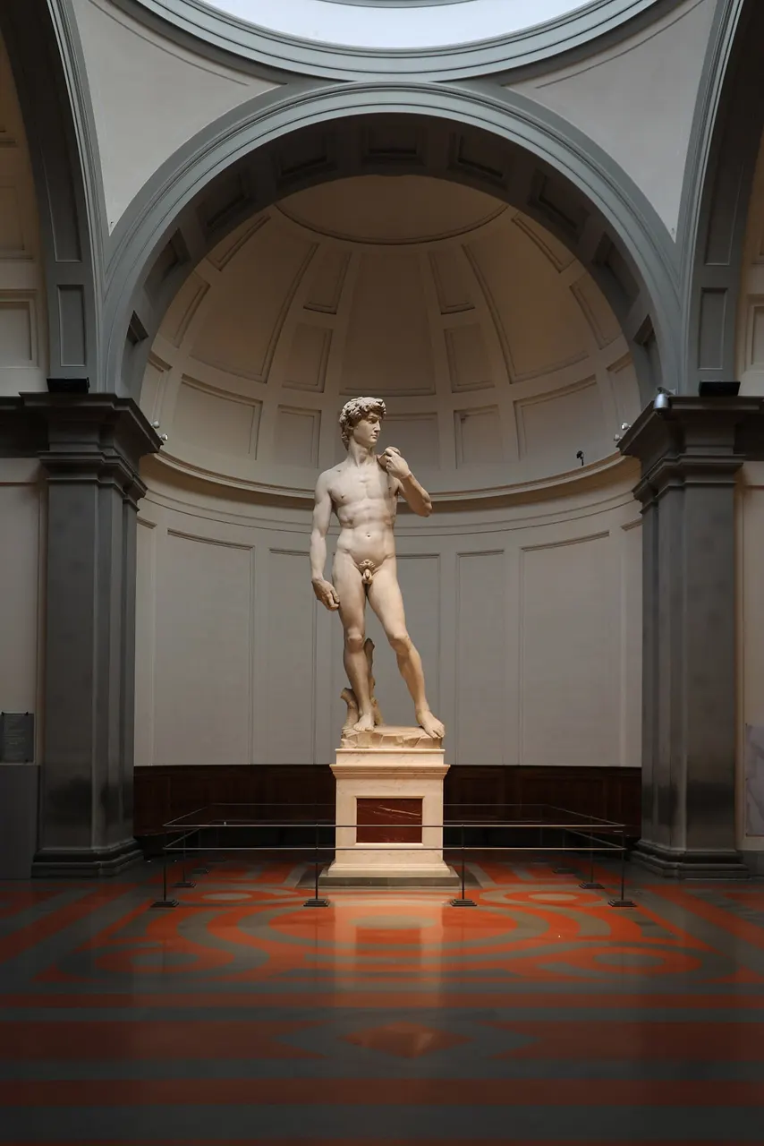 Statue de marbre blanc d’un jeune homme nu debout sur un piédestal, sous une coupole claire.