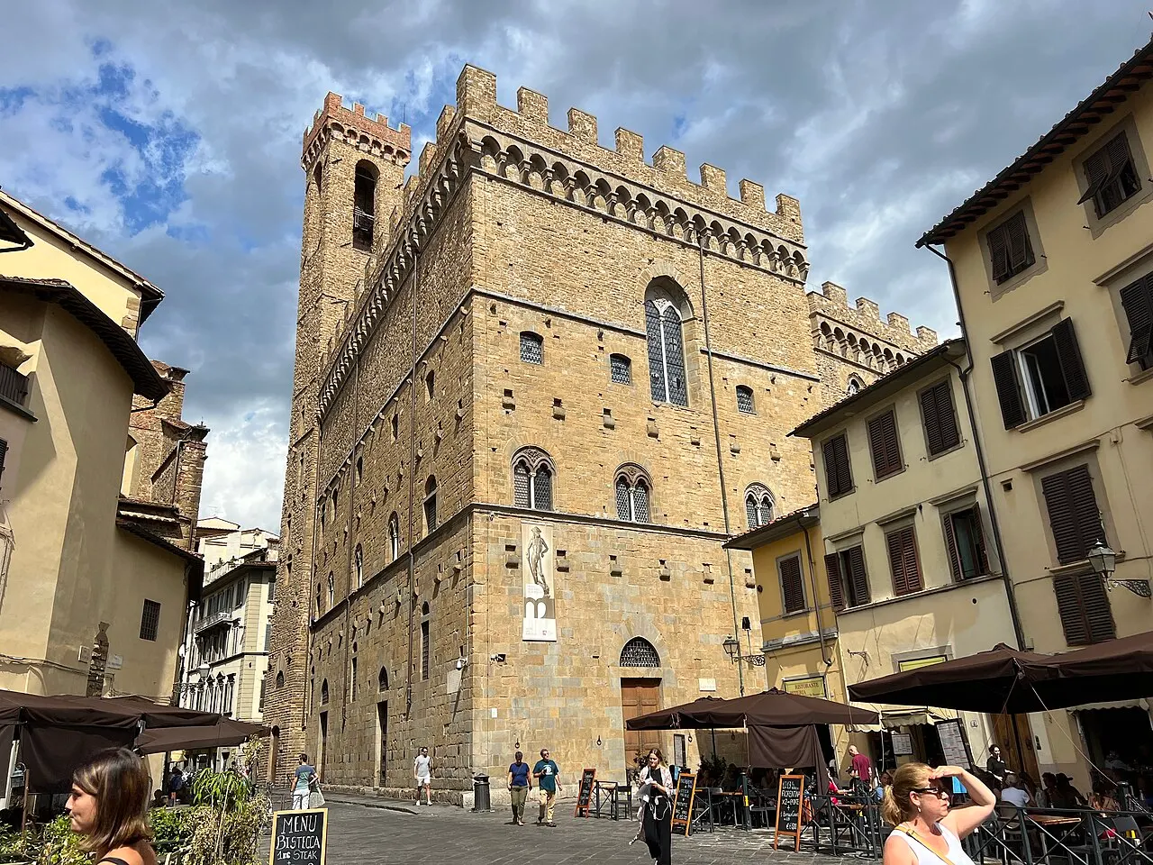 Le bloc de pierre crénelé du palais du Bargello et la tour Volognana, vus de la rue, avec des parasols de café et des passants au premier plan.