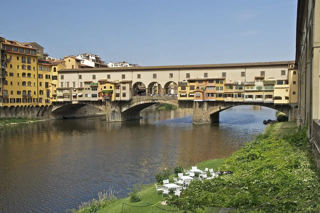Ponte Vecchio sobre o Arno, com lojas no tabuleiro, a galeria fechada por cima e o barranco verde na margem.