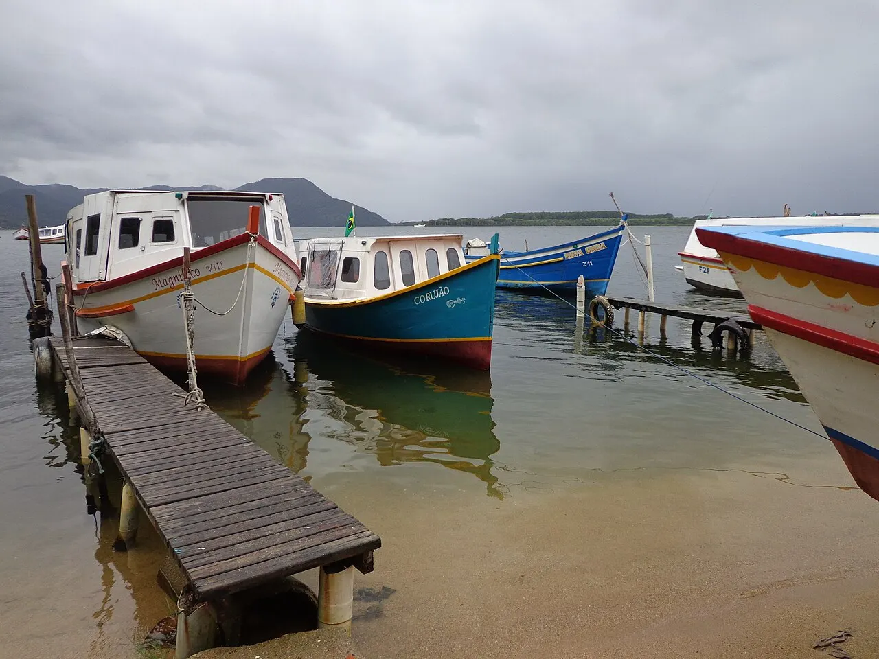 Des bateaux en bois peints en blanc, bleu et rouge amarrés à un ponton étroit, dans l’eau peu profonde de Costa da Lagoa, sous un ciel nuageux.