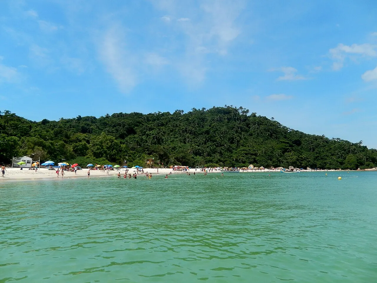 La plage de l’île de Campeche vue depuis l’eau : une mer vert clair et transparente, une bande de sable avec des parasols et des baigneurs, et la forêt sur la colline.