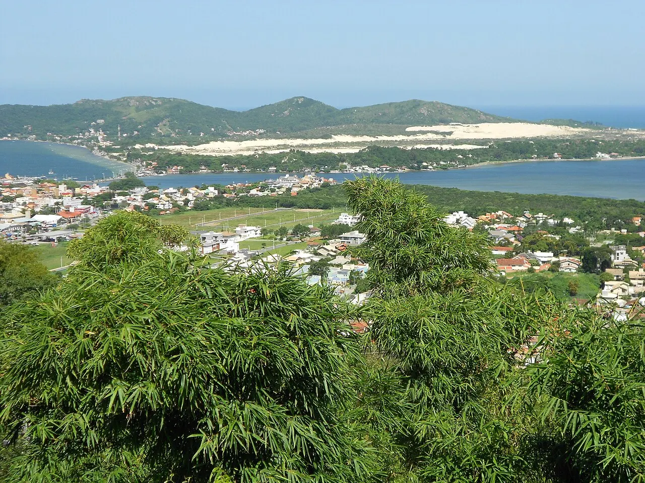 Vista do alto da Lagoa da Conceição, com casas entre a mata em primeiro plano e, do outro lado da água, as dunas claras e os morros verdes.