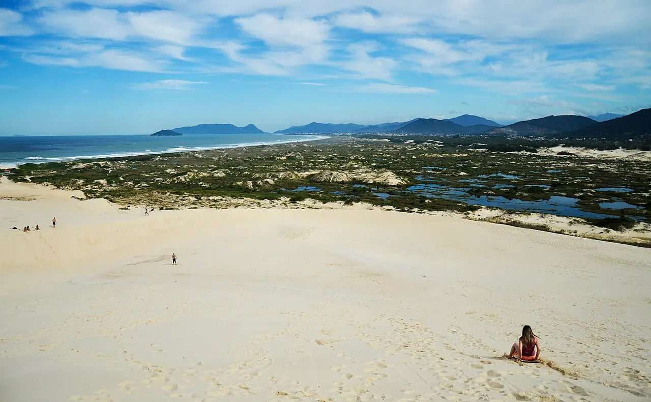 Une pente de sable clair dans les dunes de la lagune de Conceição, avec une personne assise en bas, la végétation basse du littoral et des mares plus loin, la mer et les collines à l’horizon.