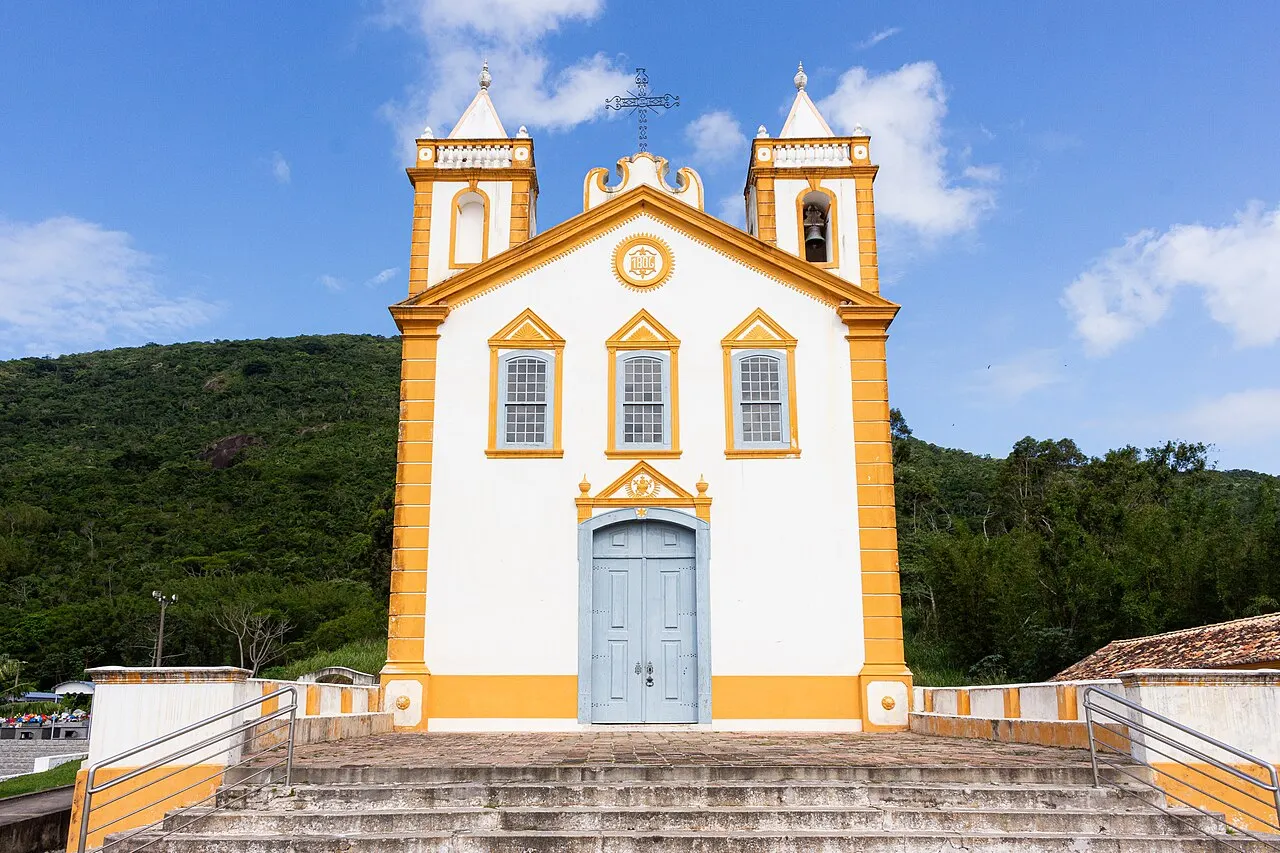 L’église Nossa Senhora da Lapa, à Ribeirão da Ilha, avec une façade blanche soulignée de jaune, deux petites tours et un large escalier, devant une colline boisée.