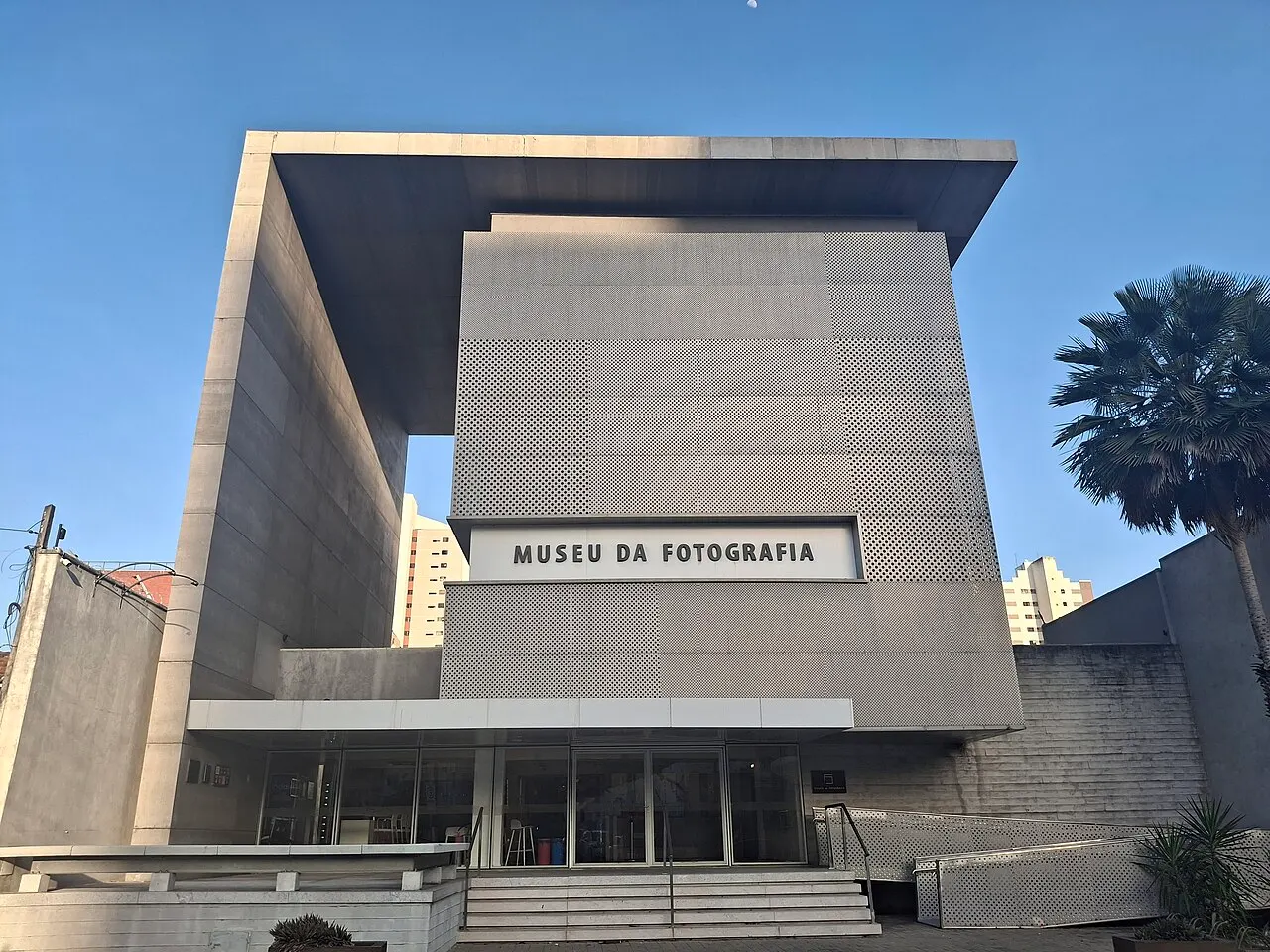 O prédio de concreto cinza do Museu da Fotografia Fortaleza, com o nome na fachada e uma palmeira ao lado.