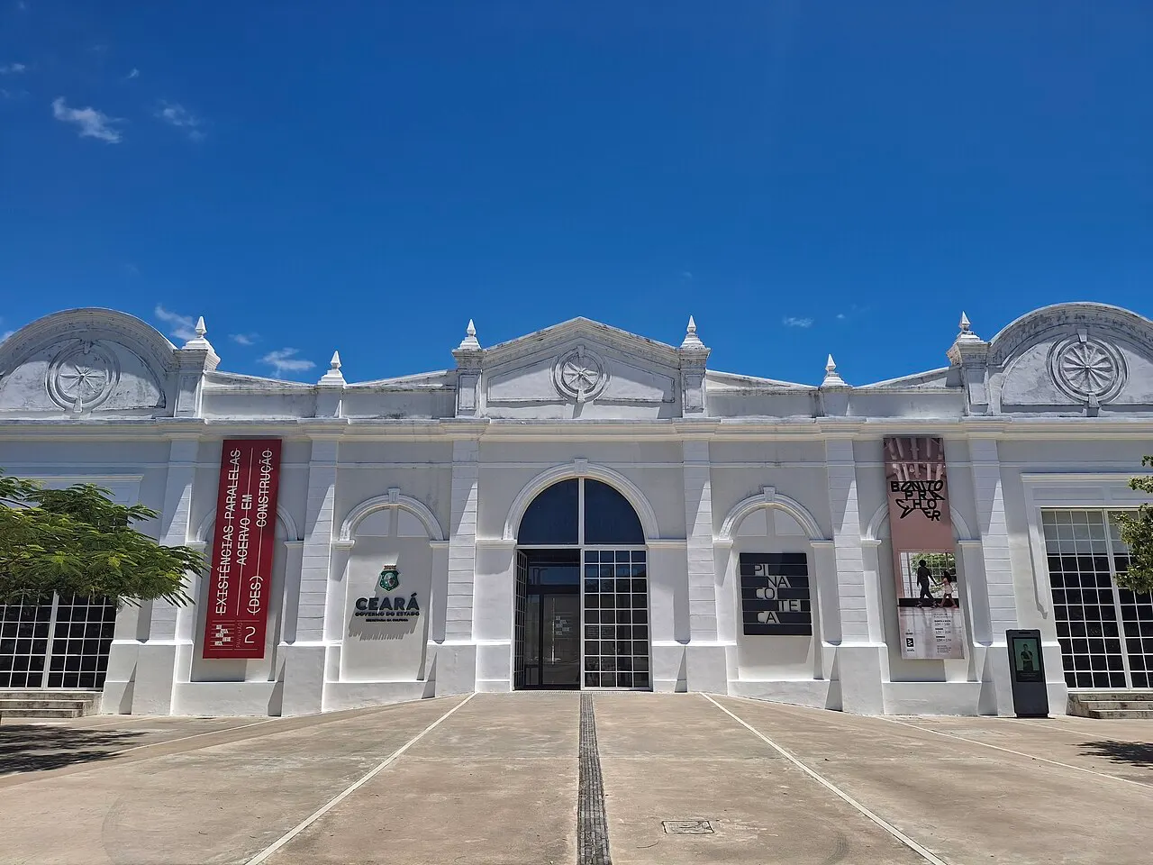 A fachada branca da Pinacoteca do Ceará, um antigo galpão ferroviário, com faixas de exposição penduradas e o pátio na frente.