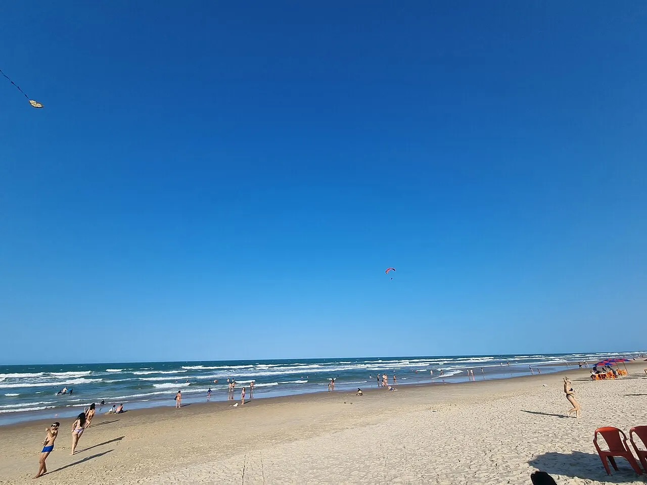 A faixa larga de areia clara da Praia do Futuro, com o mar de ondas baixas e poucas pessoas sob céu azul limpo.